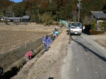 溝さらいの様子の写真で、溝の土砂をすくう人たちの奥に、ユンボ2台とトラック1台が写っている