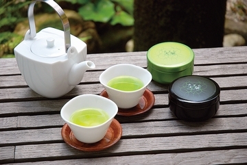 急須、緑茶が入った２つの湯飲み、黄緑と黒の入れ物が並べて置かれている写真。