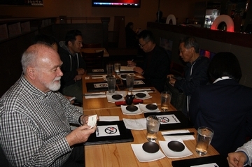 机を挟んで、6人が食事をしながら、打ち合わせをしている写真。