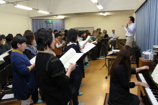 指揮者を前に合唱の練習をするたくさんの男性と女性の様子。