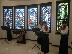 ステンドグラスの窓の前で、バイオリンやチェロ等で演奏をする5人の女性たちの写真。