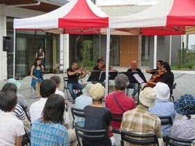 建物の外のテントの下で、座ってバイオリン等を演奏する人たちとその演奏を聴く観客の写真。
