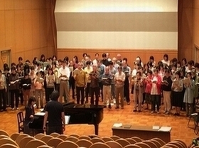 ホールで合唱の練習をする大勢の人たちの写真。手前にピアノ伴奏者と指揮者が写っている。