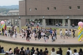 建物の前の屋外の舞台で、ダンスをする大勢の子供たちと、観客の写真。