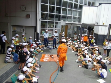オレンジ色のつなぎを着た消防隊員の説明をコの字型に座って聞いている児童らの様子
