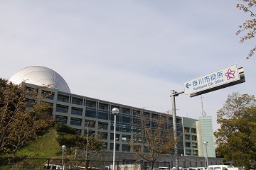 掛川市役所の道路標識とガラス張りの市役所本庁舎の写真。屋上にはドーム状の屋根が見える。