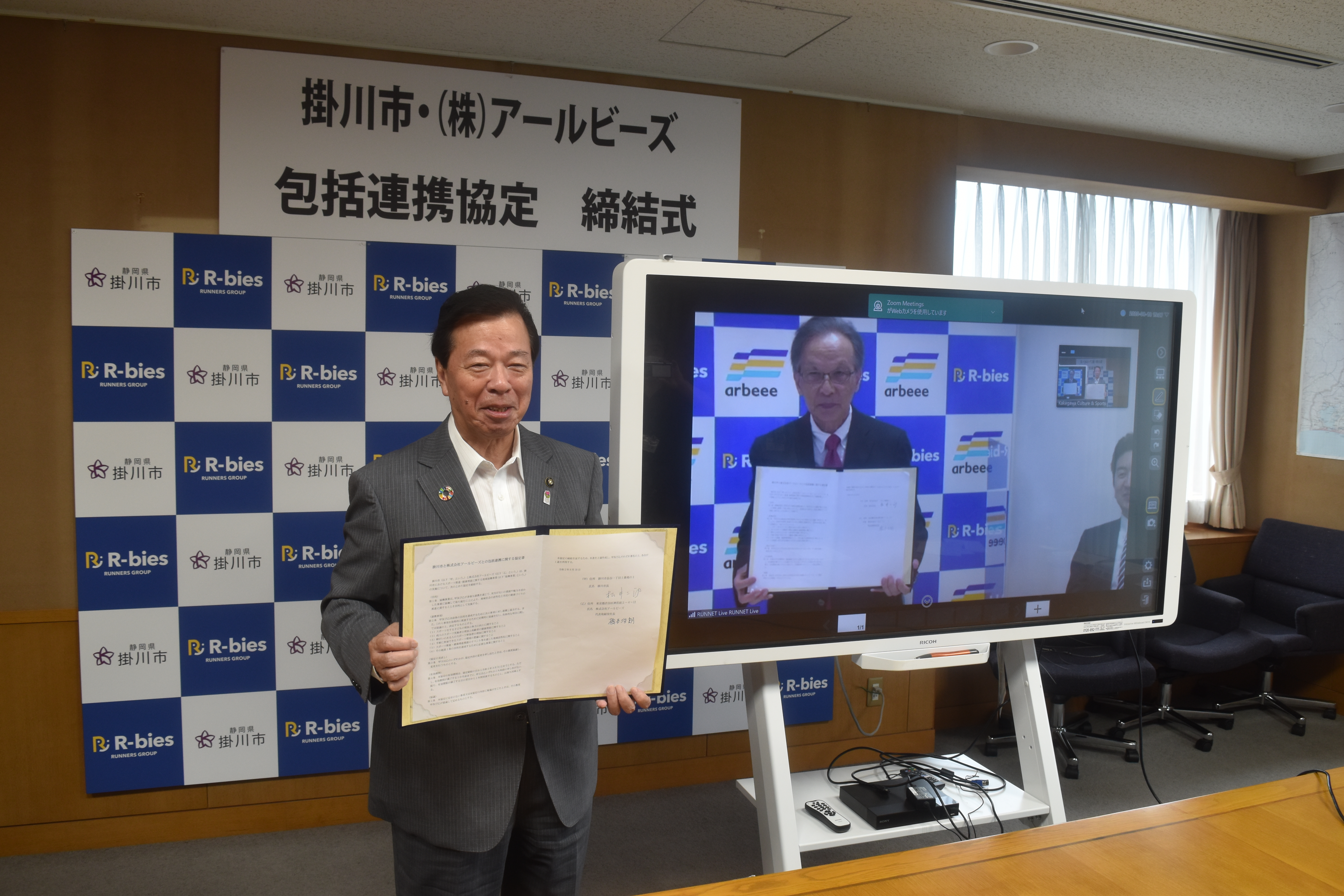 締結証書を見せる松井市長とリモートで画面の中で締結書を見せる橋本社長の写真