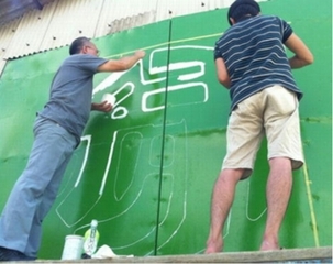 緑色に塗った屋台小屋の扉に白色のペンキで文字をデザインしているようす