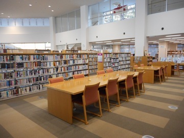 図書館の一角の写真。写真中央の本棚の前に机といすが整列している。