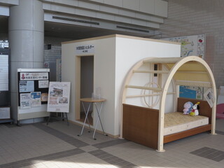 市役所ロビーに展示されている木質耐震シェルターと防災ベッドの画像で、ベッドはアーチ状のフレームがついている