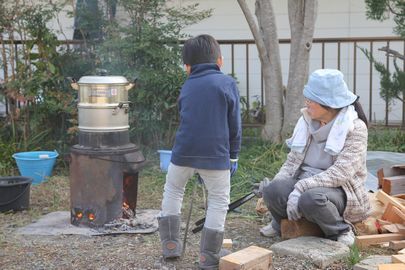 セイロで新米を蒸している様子。火箸を持ち、近くで座って火の番をしている大人と幼児。