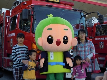 消防車の前で、茶のみやきんじろう君を挟んで、記念写真を撮っているお母さんと子供3人の写真。