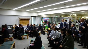 大型の映像装置の前にたくさんの人が集まっている。画面に近い方は椅子に座って、後ろの方は立ち姿勢で試合を見届ける様子