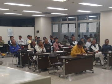 会議室。机を並べて、椅子に座って沢山の大人の人が会議をしている