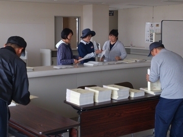 部屋で大人の人が手前に2人、奥に3人、冊子を作っている