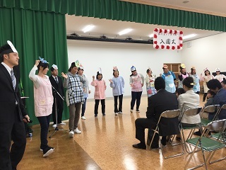 お面を頭にした職員が前に並んでポーズをしているのを子供を膝にのせパイプ椅子に座り見ている保護者たち