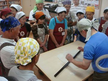 蕎麦打ちの手本を見せる人と周りを取り囲み熱心に見ている参加者たち