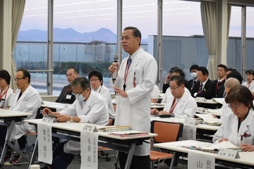 沢山の白衣を着た人らが椅子に座り、1人の白衣を着た男性がマイクを持ち、話している写真