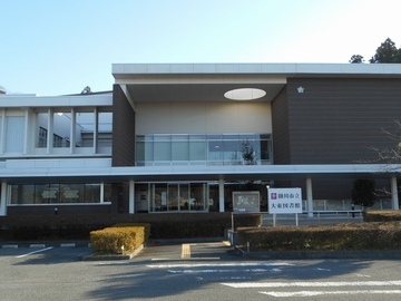 図書館の入り口付近を正面から撮った写真。手前に横断歩道、前には入り口の斜め前には駐車場が写っている。
