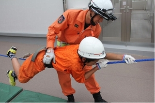 子どもが横に貼られたロープを左から右へつたう練習を、消防隊員が手助けするちびっこレスキュー体験の画像