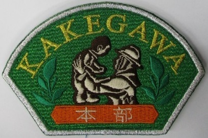 掛川市消防団の新エンブレム、変形扇形で緑地に黄色文字刺繍のKAKEGAWA、中央には消防団員が子供を抱き上げている。その左右に緑色の葉の刺繍、その下にオレンジ地に白文字刺繍の本部