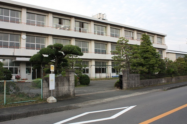 道路に面した校門の奥に三階建ての白い校舎があり、校門の手前にはバス停がある