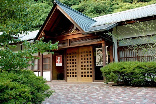 吉行淳之介文学館をほぼ正面から撮った写真。入り口にはポスターが貼ってあり、また入り口付近には緑の植木がある。