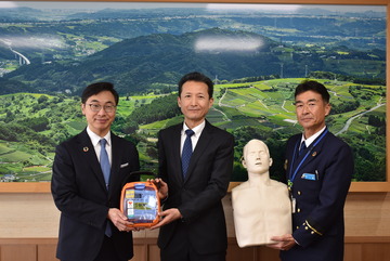 ▲久保田市長（左）にAEDトレーナーを手渡す内藤さん（中央）訓練用人形を持つ消防長（右）