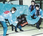 大人の人2人が獅子頭に付いている青い布の中に入り獅子舞をしている。その獅子舞の周りに3人の男性が祭りのはっぴを着て補助している
