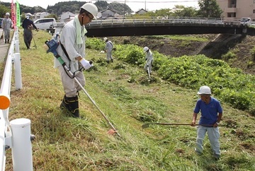ヘルメットを被り倉真川堤防の草を草刈り機で刈る会員ら