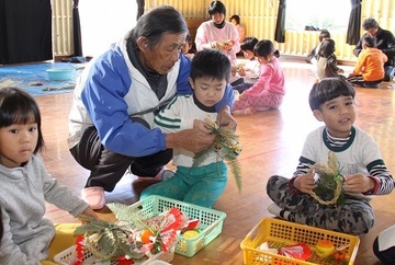 協議会のメンバーと一緒に締め飾りをもって作り方の指導をうける園児