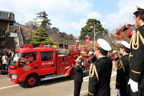 掛川城前の道路を団員やポンプ車が観閲行進し、道路脇では正装した消防団員達がラッパを吹いている