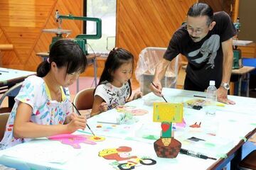 絵筆を持って指導する赤堀さんと女子児童二人