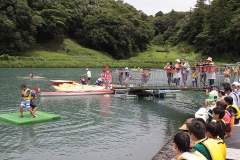 水に浮かんだマットの上で尻相撲をとる2人の男の子を池の周りでみている観客