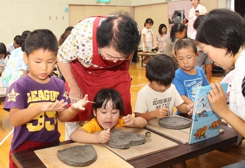 陶芸クラブの会員からお皿の飾りつけを教わる4人の園児たち。女の子がハート型のお皿に模様を描いている写真