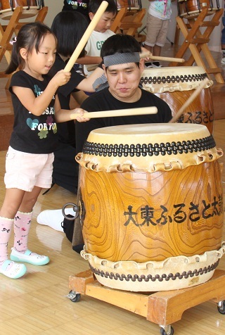 「大東ふるさと太鼓」のメンバーにアドバイスを聞きながら一生懸命に和太鼓をたたいている女子園児の写真