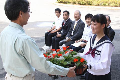 寄贈式で竹林センター副場長から赤い花の苗を受け取る鈴木さんとそれを見ている参加者
