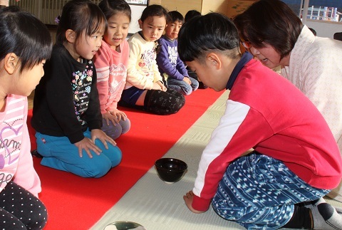 赤い毛氈の上で正座をして山田さんから茶道の礼儀作法を学ぶ園児たちの写真