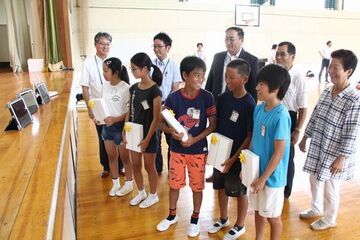 包装された箱を抱え並ぶ5人の児童と大人5人