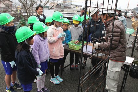 集まって鉄筋に針金を巻き付ける道具の説明を受ける緑色のヘルメットをかぶった児童たちの写真