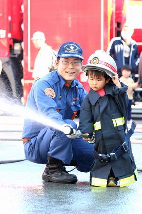 消防服を着用した子供が消防団員に支えられながら放水している