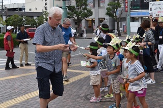 お面の様な帽子をかぶり園児らが手作りの折り紙を手渡す