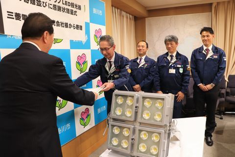 三菱電機照明深瀬工場長に感謝状を手渡す松井市長、深瀬工場長の後ろには三菱電機照明の男性社員3名、机の上には掛川城天守閣をライトアップLED投光器4台