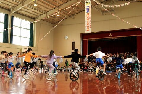 体育館の中で多くの住民を前に練習の成果を披露する児童たち、一輪車に乗り大勢の児童が手をつなぎ、大きな円になっている。