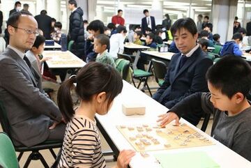 子どもたちの横に座り、対局を温かく見守る久保王将と渡辺棋王の写真