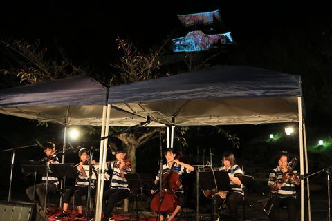 夜の掛川城と、その前で弦楽器や管楽器を演奏する高校生6人