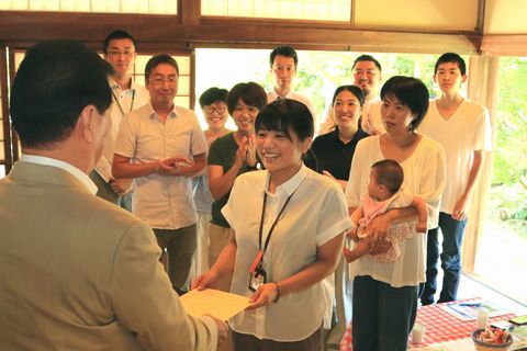 松井市長からの委嘱書を受け取る女性と、後ろ赤ちゃんを抱きながらそれを見る女性と職員たち