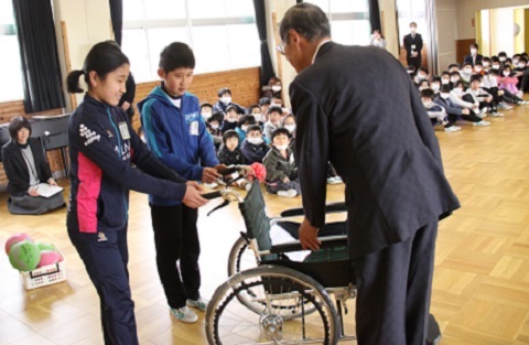 大勢の生徒の前で水野会長へ車いすを寄贈する2人の児童代表