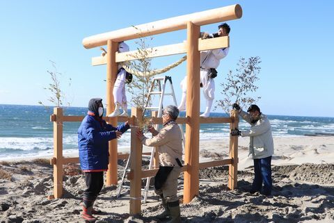 立てられた鳥居に宮大工２人が登り、しめ縄を取り付け、鳥居が倒れないように木を結び付けている3人の男の人