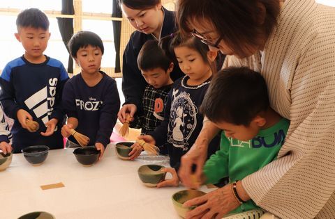 先生に手を添えられて茶筅を器で回す園児と、それをみる園児たち